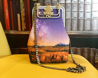 May include: A purple and gold chain bag with a landscape print. The bag has a gold clasp and a chain strap. The print features a mountain range, a field of trees, and a starry sky.