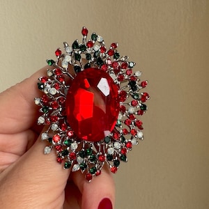 May include: A large, oval, red gemstone brooch with a silver-colored metal setting. The brooch is surrounded by smaller red, green, and white gemstones, creating a festive, floral-like design. The brooch is held by a hand with red nail polish.