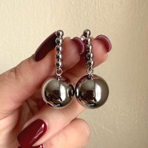 May include: A pair of silver-colored dangle earrings. Each earring features a chain of small, round beads leading to a large, reflective sphere. The earrings are shown against a neutral background.