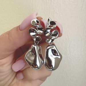 May include: A pair of silver-toned drop earrings featuring a polished, reflective finish. Each earring consists of a spiraled, ribbon-like top section connected to a smooth, irregular, organic-shaped pendant that hangs vertically from the ear wire.