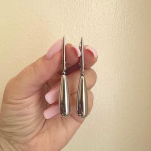May include: A pair of silver-toned drop earrings featuring smooth, elongated teardrop-shaped pendants suspended from thin metal hooks. The earrings have a polished, reflective metallic finish and a simple, clean design with no additional embellishments.