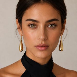 May include: A pair of polished gold-tone drop earrings featuring a smooth, elongated teardrop shape. The earrings are suspended from a simple, thin vertical bar attachment that connects to the earlobe, creating a clean and reflective metallic finish.