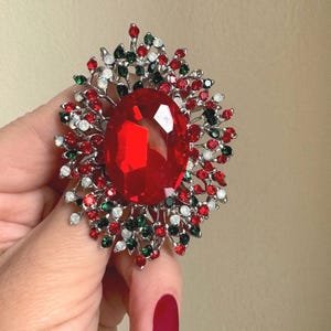 May include: A large, oval, red gemstone brooch with a silver-tone metal setting. The brooch is surrounded by smaller red, green, white, and black crystals, creating a sparkling, festive design. The brooch is held by a person.