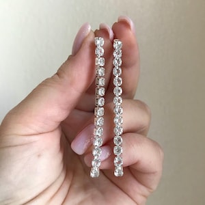 May include: Two silver-toned, linear drop earrings. Each earring features a series of clear, sparkling stones set in a vertical line. The stones are of varying shapes, some square and some round, creating a shimmering effect.