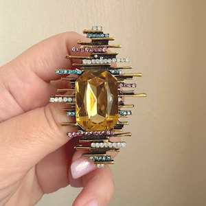 May include: A gold-toned brooch with a large, rectangular yellow gemstone at its center. The brooch is adorned with small, colorful gemstones in shades of blue, pink, and white, arranged around the central stone.