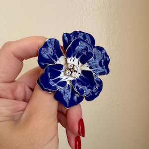May include: A large, flower-shaped brooch with a glossy, deep blue enamel finish. The petals have white accents and gold trim. The center features a cluster of small, sparkling crystals. A statement piece of jewelry.
