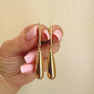 May include: A pair of polished gold-tone drop earrings featuring a smooth, elongated teardrop shape. Each earring is suspended from a thin, vertical metal post that connects to the top of the metallic, reflective, and solid teardrop pendant.