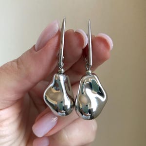 May include: A pair of silver-colored, abstract-shaped earrings. Each earring features a long, thin hook and a rounded, irregular pendant. The pendants have a reflective, polished surface, catching the light and creating highlights.