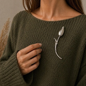 May include: A silver-colored tulip brooch pinned to a dark green sweater. The brooch is a detailed representation of a tulip flower with a curved stem. The sweater has a ribbed knit texture.