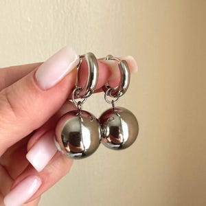 May include: A pair of silver-colored hoop earrings. Each earring features a large, reflective sphere suspended from a hinged hoop. The earrings are shown against a neutral background.