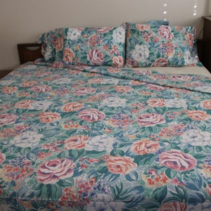 May include: A floral bed set featuring a comforter and two pillowcases. The pattern includes large pink, white, and orange roses, with smaller purple and blue flowers on a teal background. The bed set is on a wooden bed frame.