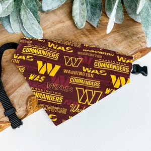 May include: A maroon and gold dog bandana featuring the text "WAS WASHINGTON COMMANDERS" and the letter "W". The bandana has a black adjustable strap and buckle. The bandana is made of fabric.