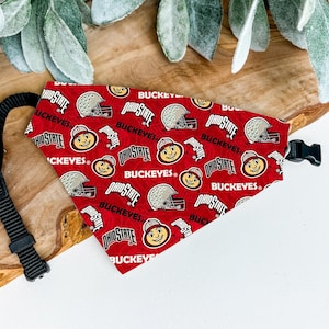 May include: Red dog bandana with a repeating pattern of Ohio State Buckeyes logos, helmets, and the word "Buckeyes." The bandana has a black adjustable strap and buckle.