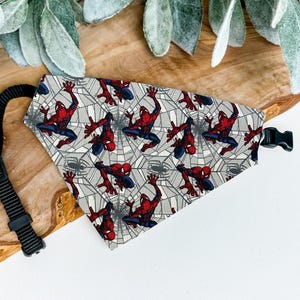 A Spiderman-themed bandana is displayed on a wooden surface, with a black leash attached to it.