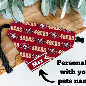 May include: A red pet bandana featuring a repeating gold and white 49ers logo pattern and the name Max printed on a white banner. The bandana includes a black adjustable nylon strap with a plastic buckle closure for secure attachment.