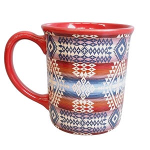 Puede incluir: Una taza de cerámica con asa y borde rojos, que presenta un patrón geométrico repetitivo en tonos azul, morado, naranja y blanco. El diseño consiste en diamantes apilados, cruces y rayas horizontales que envuelven toda la superficie exterior.