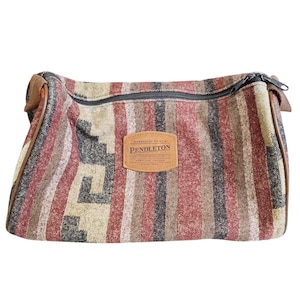 May include: A rectangular zippered pouch featuring a woven textile pattern in shades of red, tan, black, and cream. A tan leather patch is centered on the front with the text "Warranted to be a Pendleton, Pendleton Woolen Mills, Portland, Oregon, Since 1863."