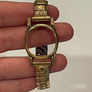 May include: A gold-tone watch case with an empty circular dial opening, featuring a flexible metal link bracelet with a textured diamond-patterned design. The watch case is polished and metallic, showing no internal watch face or hands.