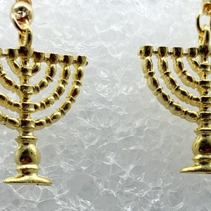May include: A pair of gold-tone earrings featuring a menorah design. The menorah is a traditional Jewish symbol of the Festival of Lights, Hanukkah.