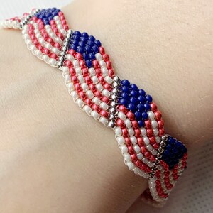May include: A beaded bracelet featuring a design reminiscent of the American flag. The bracelet is made with red, white, and blue beads, with silver-colored connectors. The design includes stripes and a field of blue with white stars.