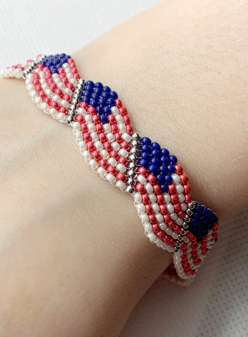 May include: A beaded bracelet featuring a design reminiscent of the American flag. The bracelet is made with red, white, and blue beads, with silver-colored connectors. The design includes stripes and a field of blue with white stars.