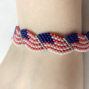 May include: A beaded bracelet featuring a design reminiscent of the American flag. The bracelet showcases alternating stripes of red and white beads, with a section of blue beads representing the stars. The bracelet is worn on a wrist.
