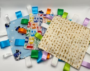 May include: Three square fabric sensory blankets with colorful satin ribbon tags. One blanket features a light blue background with cartoon characters and snowflakes, another has a multi-colored Hebrew alphabet print, and the third has a matzah pattern.