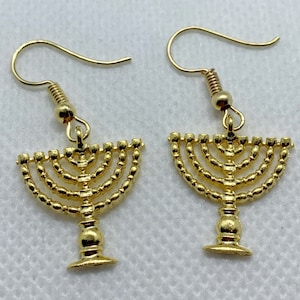 May include: A pair of gold-tone earrings featuring a menorah design. The earrings are made of metal and have a hook closure.