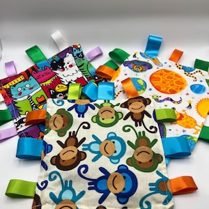May include: A collection of baby tag blankets with colorful patterns and satin ribbons. The blankets feature various designs, including cartoon cats, monkeys, and planets. Each blanket has multiple ribbons in different colors, such as green, blue, and orange.
