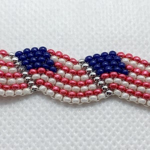 May include: A beaded bracelet or trim featuring a wavy pattern in red, white, and blue, reminiscent of the American flag. The beads are small and round, with silver accents. The design is patriotic and decorative.