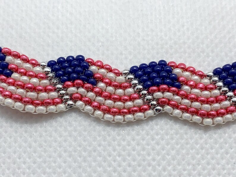 May include: A beaded bracelet or trim featuring a wavy pattern in red, white, and blue, reminiscent of the American flag. The beads are small and round, with silver accents. The design is patriotic and decorative.