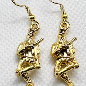 Może przedstawiać: Para złotych kolczyków z detaliczną figurką osoby trzymającej karabin. Kolczyki mają zapięcie na haczyk.
