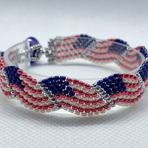 May include: A beaded bracelet featuring a patriotic design with red, white, and blue beads. The bracelet is made with small, tightly woven beads, creating a textured surface. The clasp is silver-toned, and the bracelet is displayed on a clear, curved form.