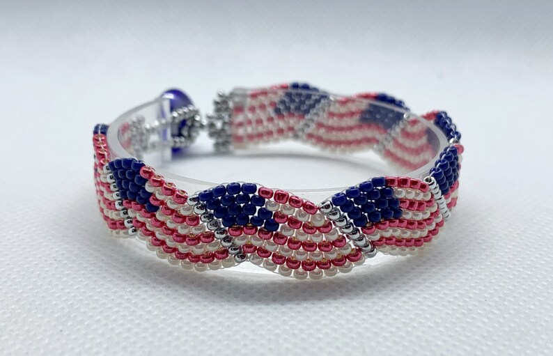 May include: A beaded bracelet featuring a patriotic design with red, white, and blue beads. The bracelet is made with small, tightly woven beads, creating a textured surface. The clasp is silver-toned, and the bracelet is displayed on a clear, curved form.