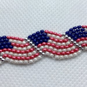 May include: A beaded bracelet featuring a design reminiscent of the American flag. The bracelet showcases alternating rows of red, white, and blue beads, with a silver beaded border. The design is a wavy pattern.