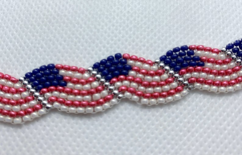 May include: A beaded bracelet featuring a design reminiscent of the American flag. The bracelet showcases alternating rows of red, white, and blue beads, with a silver beaded border. The design is a wavy pattern.