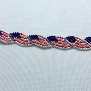 May include: A beaded bracelet featuring a repeating pattern of American flags. The bracelet is made with red, white, and blue beads, with a silver-colored chain and a blue button clasp. The bracelet is approximately 7 inches long.