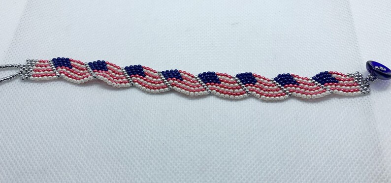 May include: A beaded bracelet featuring a repeating pattern of American flags. The bracelet is made with red, white, and blue beads, with a silver-colored chain and a blue button clasp. The bracelet is approximately 7 inches long.