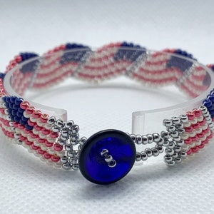 May include: A beaded bracelet featuring a design reminiscent of the American flag. The bracelet showcases alternating stripes of red and white beads, with a blue section adorned with white beads. A dark blue button serves as the clasp.