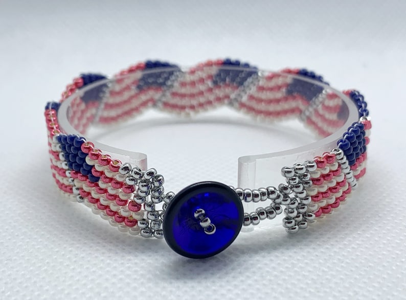 May include: A beaded bracelet featuring a design reminiscent of the American flag. The bracelet showcases alternating stripes of red and white beads, with a blue section adorned with white beads. A dark blue button serves as the clasp.