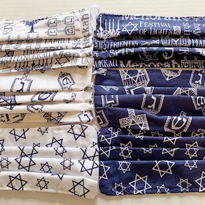 Puede incluir: Conjunto de seis mascarillas faciales con un tema de Hanukkah en azul y blanco. Las mascarillas tienen una variedad de diseños que incluyen estrellas de David, dreidels, menorahs y las palabras hebreas "Feliz Janucá".