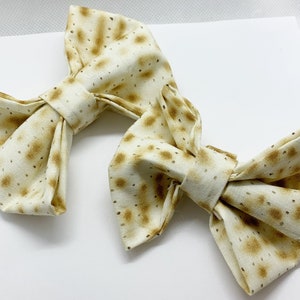 May include: Two fabric bow ties with a matzah pattern. The bows are white with brown spots.