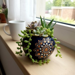 Può includere: Una fioriera in ceramica blu navy con un motivo a pois dipinto a mano in oro e bianco, piena di una varietà di piante grasse. Una pianta verde a collana di perle ricade sul lato. Una tazza bianca è vicina.