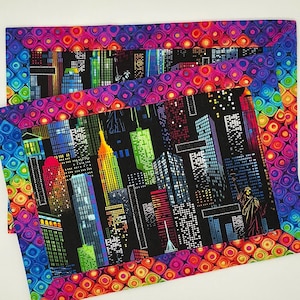 Könnte beinhalten: Zwei farbenfrohe Tischsets mit einem Stadtbild-Design. Die Tischsets haben einen schwarzen Hintergrund mit einem regenbogenfarbenen Stadtbild-Design und einem mehrfarbigen Rand.