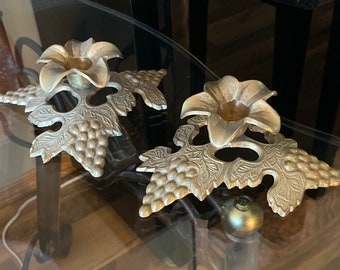 May include: Two gold metal candle holders with a grapevine design. Each holder has a flower-shaped top with a small cup for a candle.