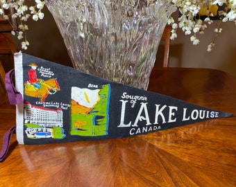 Könnte beinhalten: Schwarzer Filz-Wimpel mit weißem Text, der "Souvenir of LAKE LOUISE CANADA" lautet. Der Wimpel zeigt bunte Illustrationen eines Mounties auf dem Pferd, eines Hotels und einer Bergszene.