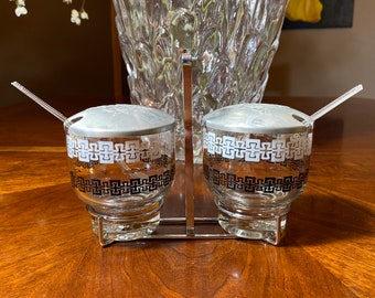 May include: A set of two glass salt and pepper shakers with black and white geometric designs. The shakers are on a silver stand with matching spoons.