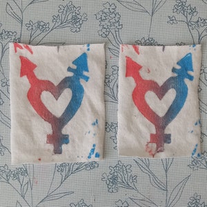 May include: Two white fabric squares with a red and blue gender symbol and heart design printed on them. The symbol is a combination of the male and female symbols.