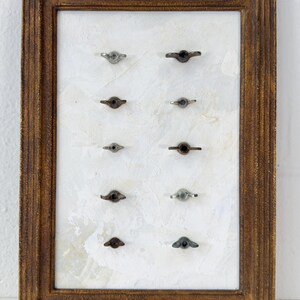 May include: A framed display of vintage metal wing nuts. The wing nuts are arranged in rows on a textured white background. The frame is made of dark brown wood with a distressed finish, adding a rustic aesthetic.