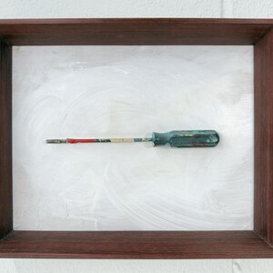 May include: A framed artwork featuring a vintage screwdriver. The screwdriver has a blue handle and a metal shaft with red and white paint. The frame is a dark brown colour, and the background is white with brushstrokes.